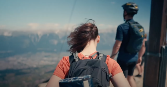Junge Frau mit Rucksack und junger Mann mit Helm sind von hinten zu sehen und schauen in Richtung der Tiroler Berge.