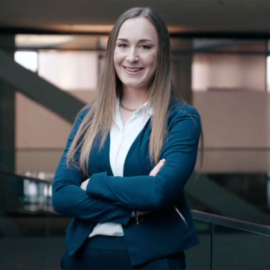Eine junge Frau im Business-Outfit steht selbstbewusst mit verschränkten Armen in einem modernen Bürogebäude.